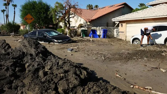 Mud and storm debris piled along a residential street with palms and tile-roof homes after a winter storm
